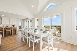 A spacious dining area with a long table, white chairs, and a view of the outdoors through large windows.
