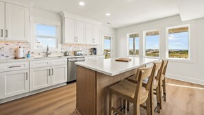 A modern kitchen with white cabinets, a central island with bar stools, and large windows offering a view of the landscape. - NEWLY RENOVATED! Oceanfront, Beach Access, Dog Friendly, Spacious Decks (Salvo)