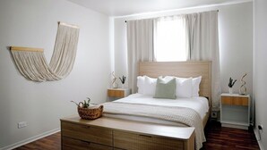 Ein Schlafzimmer mit einem hölzernen Bett, weißer Bettwäsche, einem Nachttisch mit Pflanze, einer hängenden Makramee-Wanddekoration und einer Holzkommode.