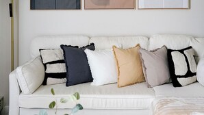 Un canapé blanc agrémenté de plusieurs coussins de différentes couleurs et motifs, un lampadaire et trois œuvres d’art abstraites encadrées accrochées au mur.