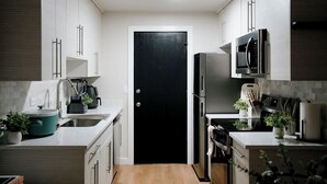 Une cuisine moderne avec des meubles blancs, une porte noire, des appareils électroménagers en acier inoxydable et un petit plan de travail sur lequel se trouvent une bouilloire et une plante.