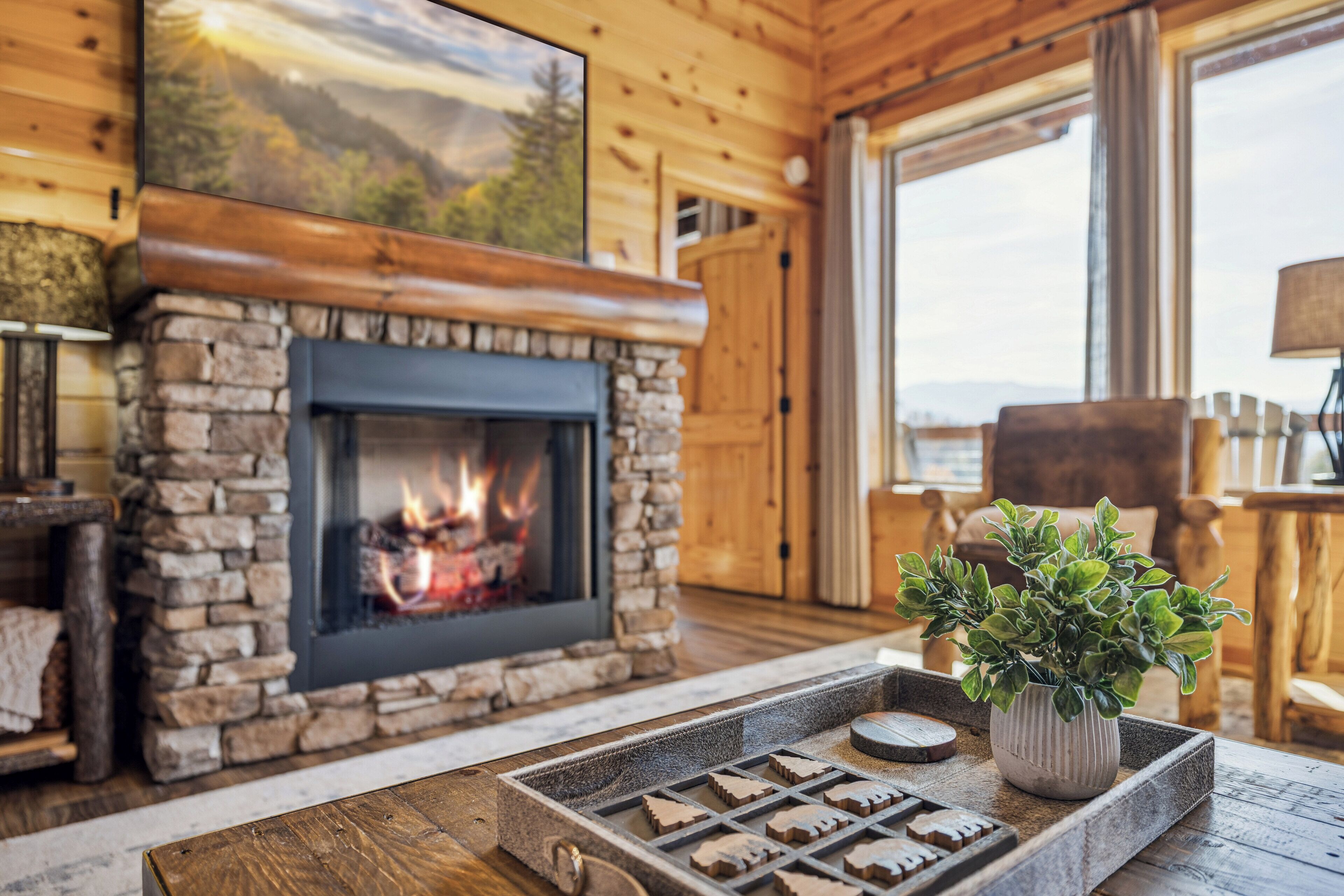 A cozy cabin interior with a stone fireplace, a wooden ceiling, and a large window.