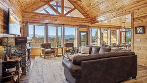En stuga med träpanel på insidan, en steneldstad, en stor soffa och en matplats med utsikt över bergen.