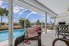 A covered outdoor area with a pool, a chair with a striped towel, and a table with a grill. - Serene Canal Views, Heated Pool & Relaxation - Villa Pelican Paradise - Roelens (Cape Coral)