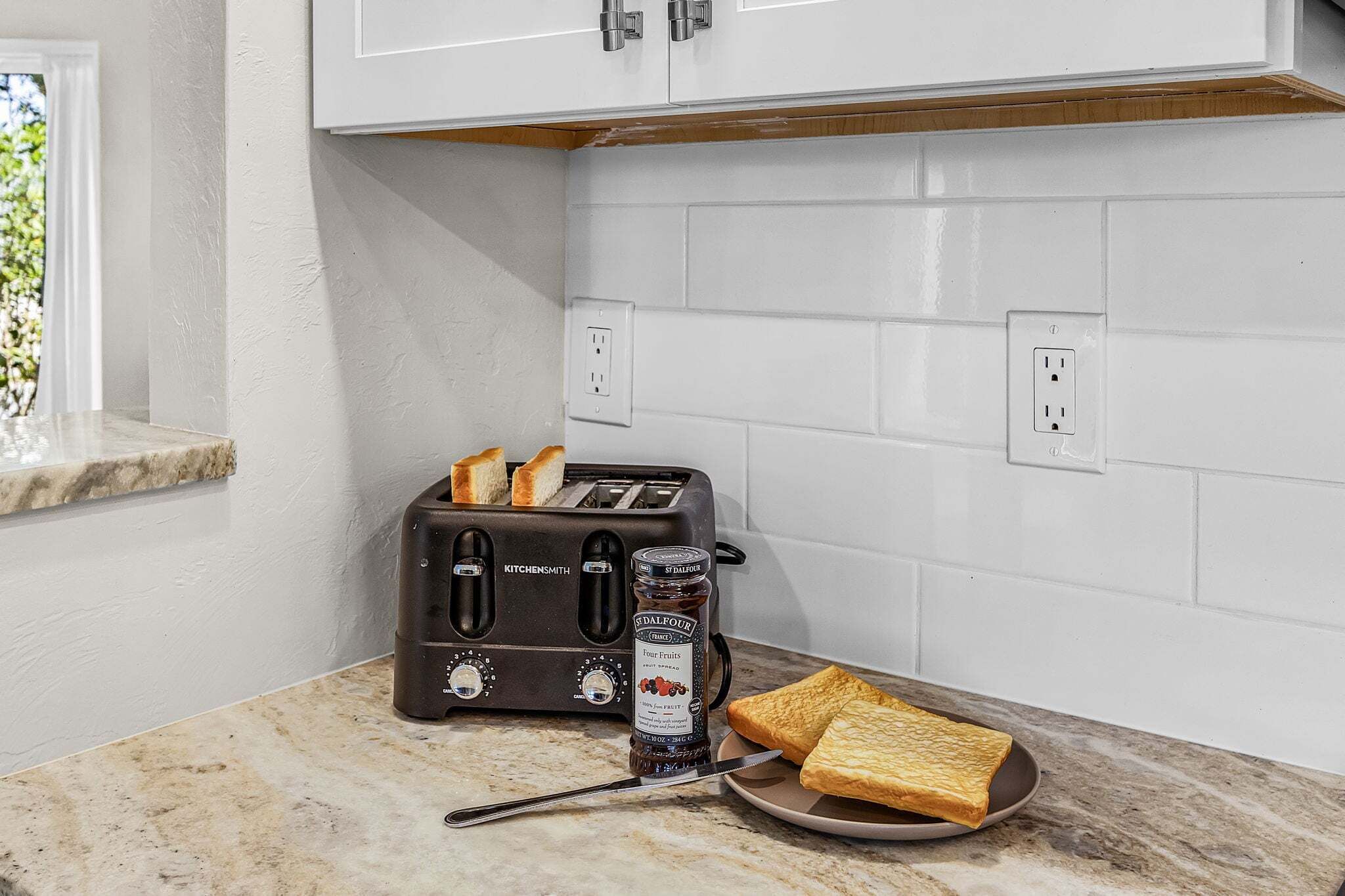 Een keuken met een broodrooster, een pot jam en twee sneetjes toast op een marmeren aanrecht.
