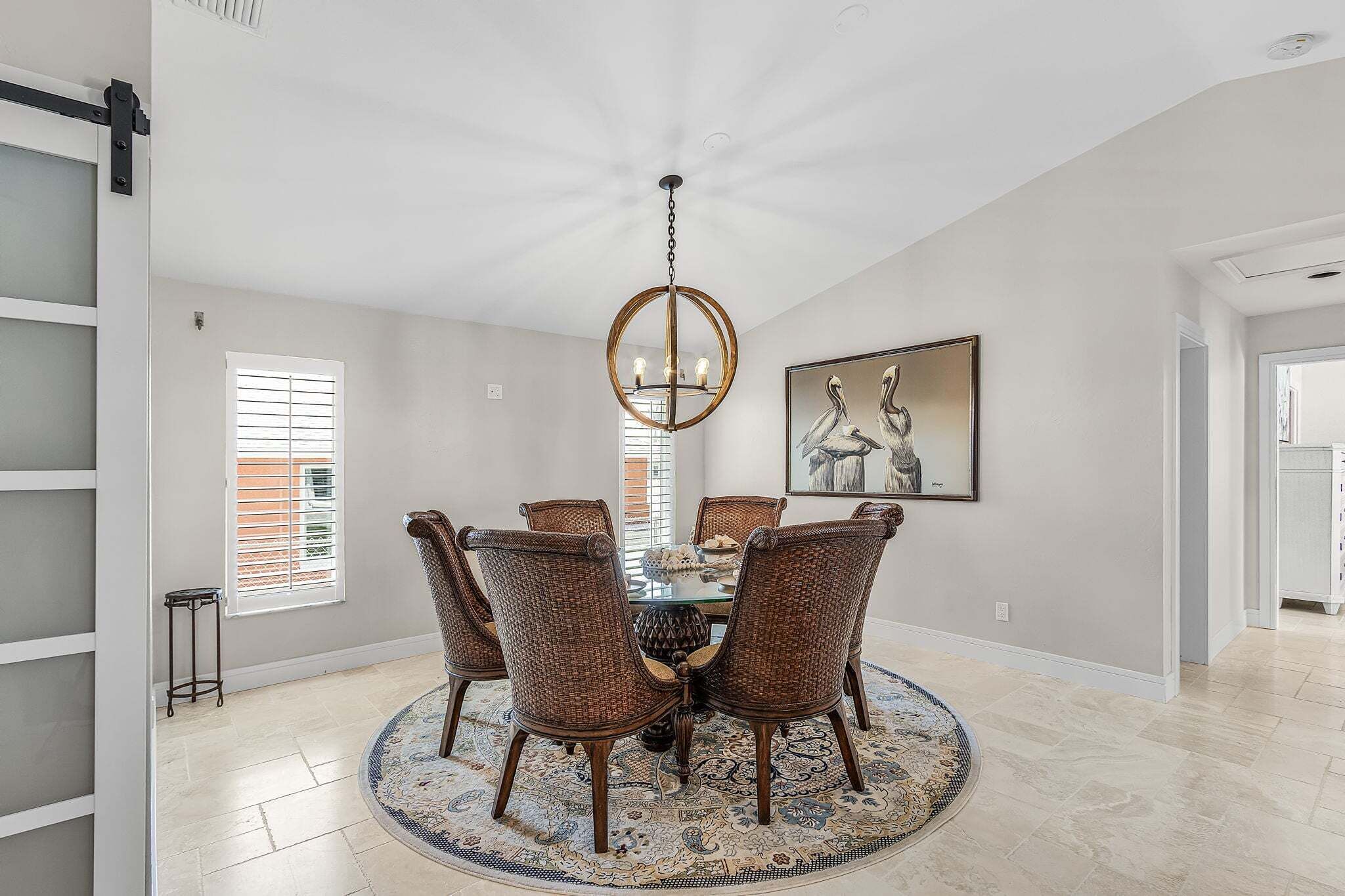 A dining room with a round table, six wicker chairs, a chandelier, a painting, and a patterned rug.