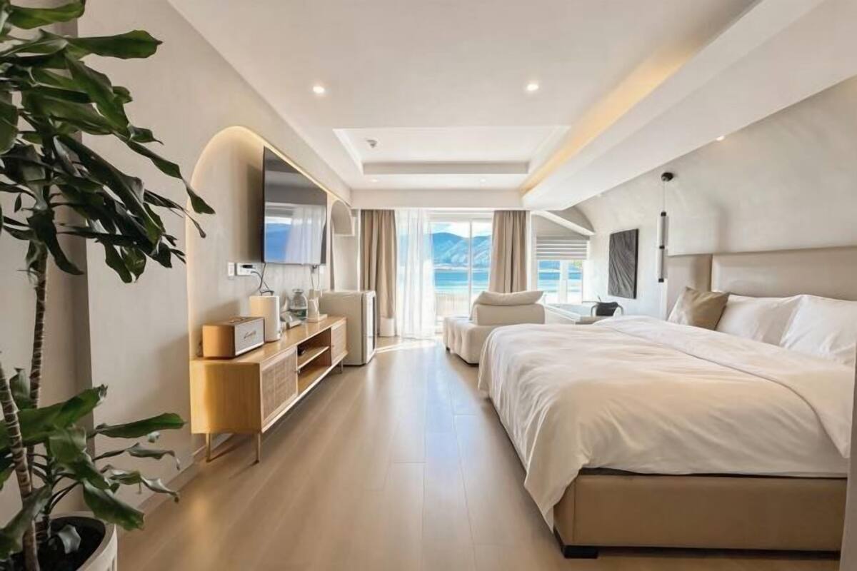 A modern hotel room with a large bed, a TV, a desk, and a view of the sea.