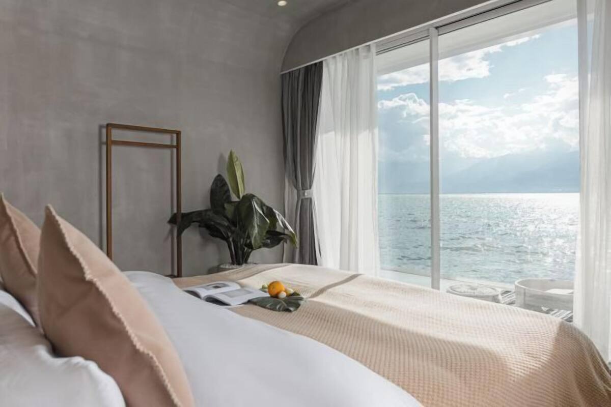 A bedroom with a large window overlooking a body of water, a bed with pillows, a bedside table with a book and fruit, and a plant.