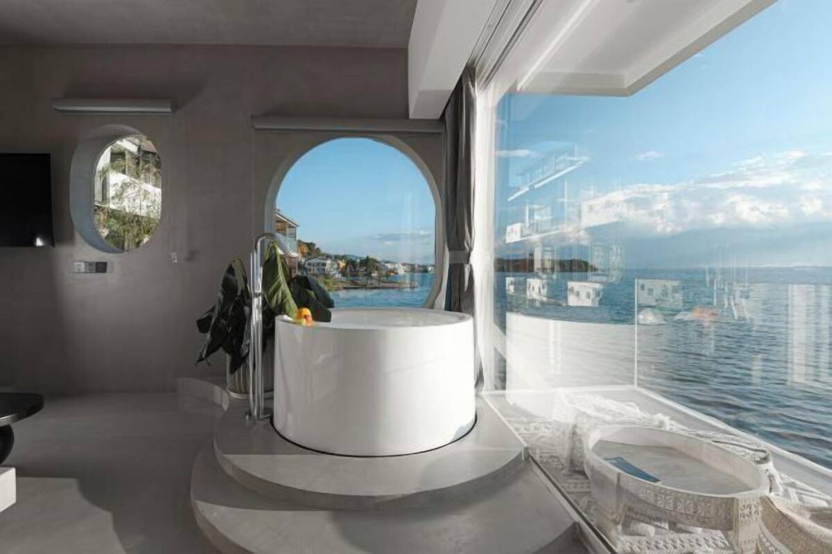 A modern bathroom with a circular sink, a large window with a view of the sea, and a small circular window.