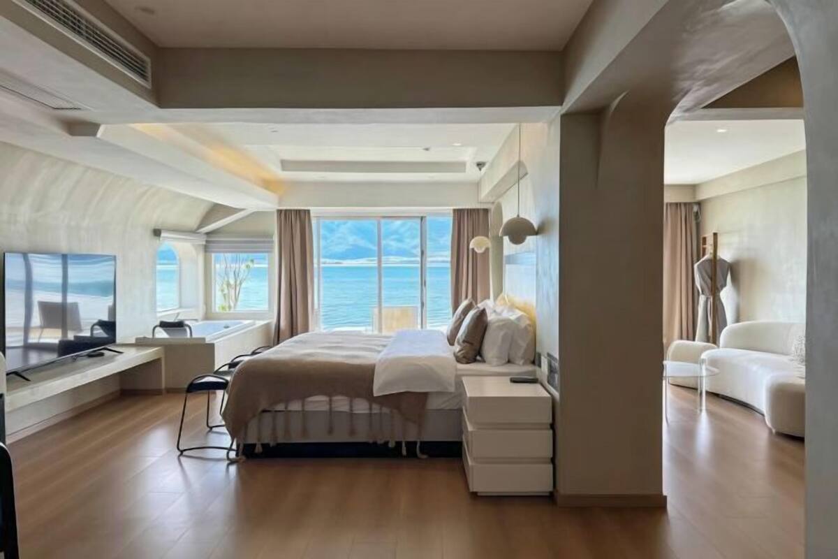 A spacious bedroom with a large bed, a desk, and a TV. There is a view of the sea through large windows.