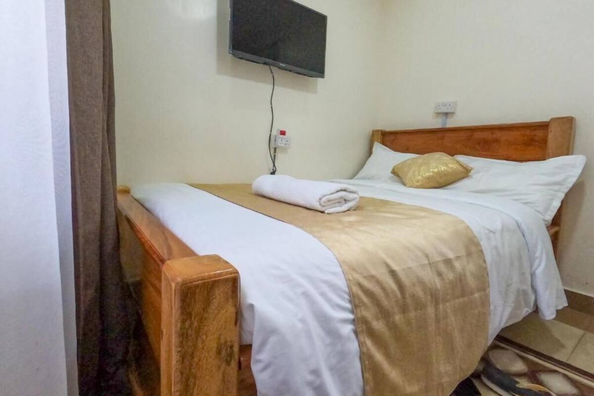 Un lit simple avec une tête de lit en bois, une literie blanche et une couverture beige. Un téléviseur est fixé au mur au-dessus du lit.