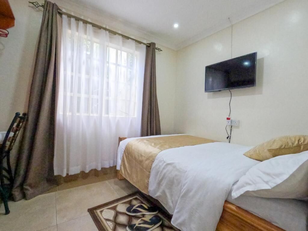 Ein Hotelzimmer mit einem Bett, einem an der Wand befestigten Fernseher, einem Fenster mit Vorhängen und einem Paar Hausschuhen auf dem Boden.