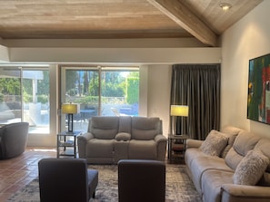 Un salon avec un canapé beige, un fauteuil inclinable et une table basse. Il y a une grande fenêtre avec des rideaux, un tableau au mur et une lampe sur une table d’appoint.