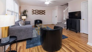 Un salon avec un canapé gris, un pouf noir, une chaise noire, un téléviseur posé sur un meuble noir et un escalier en arrière-plan.