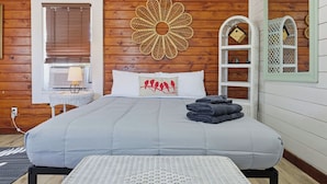 Ein gemütliches Schlafzimmer mit einem großen Bett, einer Holzwand, Wandkunst mit Blättern, einem kleinen Nachttisch mit Lampe und einem weißen Korbstuhl.