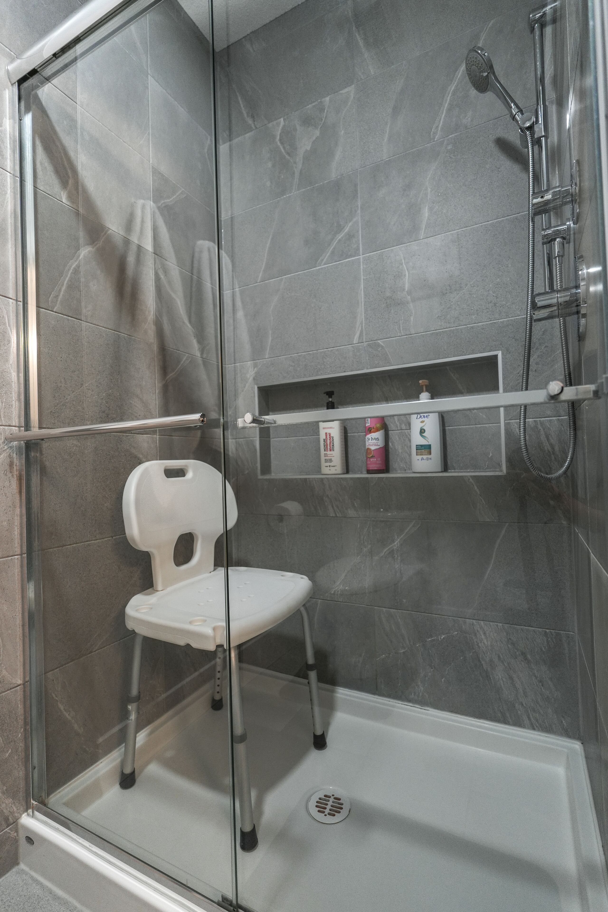 A shower area with a glass door, a white chair, and a shelf with toiletries.