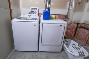 Dos lavadoras blancas, un recipiente azul para agua, una caja de detergente verde y una cesta de ropa blanca.