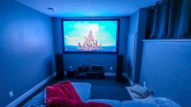 A room with a large TV screen displaying a castle, a bed, a red blanket, and speakers. - 4BR Dream Family Stay! ☆ King Bed / Home Theatre / Large Backyard ☆ Pet Friendly (Sturgeon County)