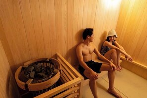 Zwei Personen sitzen auf Holzbänken in einer Sauna mit einer Holzverkleidung und einem Steinkessel.