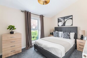A bedroom with a bed, bedside tables, a dresser, a window with curtains, and a view of buildings outside. - Cozy House w/garden near 02 Arena sleeps 10, 2 mins to station (London)