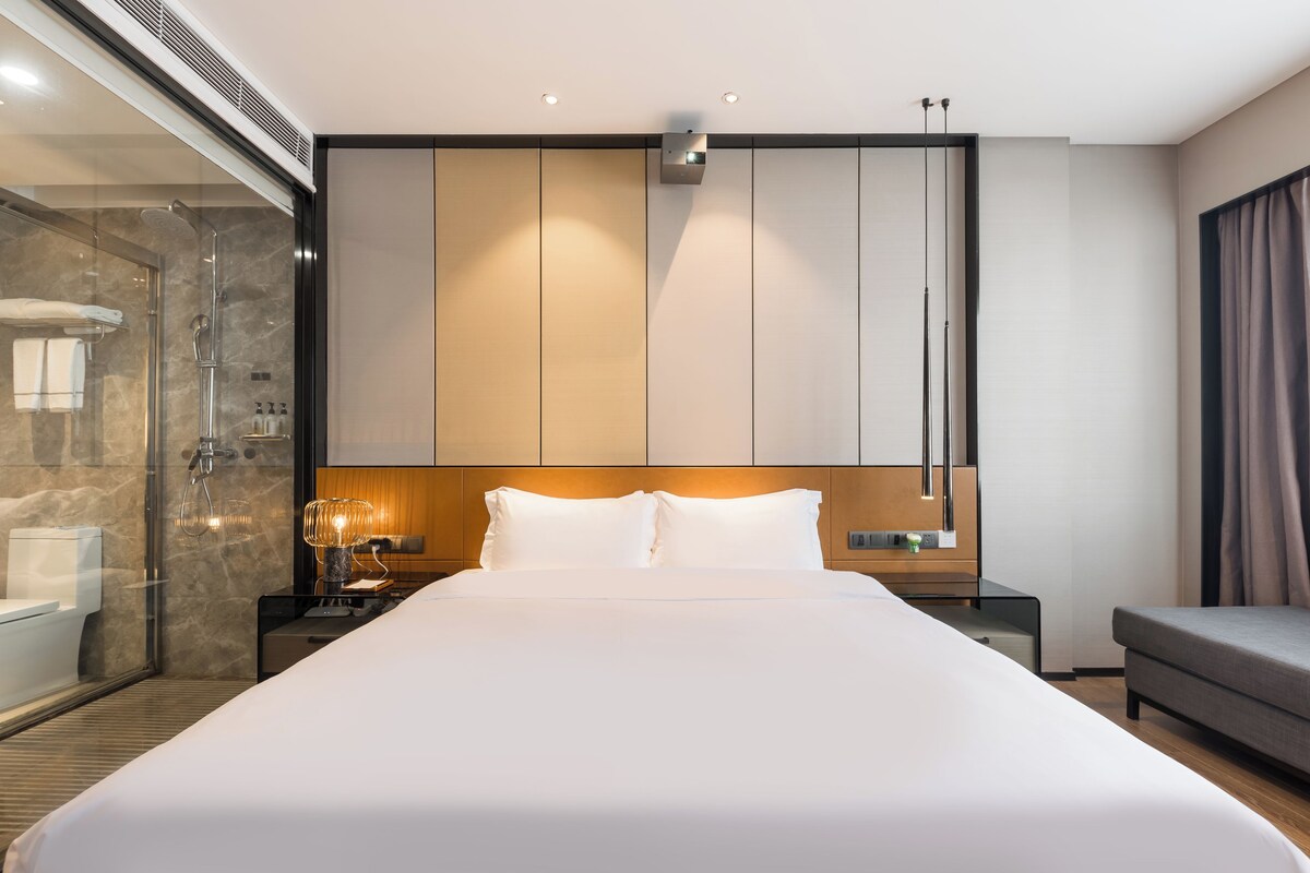 A modern hotel room with a large bed, a glass-enclosed shower, a wooden headboard, and a nightstand with a lamp.