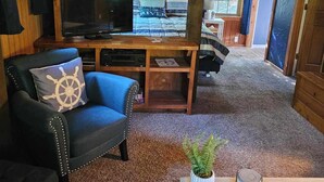 Ein gemütliches Blockhausszimmer mit Holzboden, einem Fernseher auf einem Holztisch, einem blauen Sessel mit Kopfkissen in Form eines Schiffsschwals und einem kleinen Tisch mit Pflanze und einem Schild mit der Aufschrift 'Relax'.