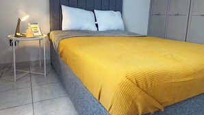 Et soveværelse med en grå sengegavl, to hvide puder, et gult tæppe og et natbord med en lampe og et billede i ramme.