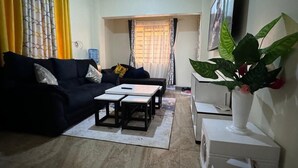 A living room with a black sofa, two small tables, a white cabinet, and a potted plant. - G&M Luxe Abode Vacation Rental Nyeri (King'ong'o)