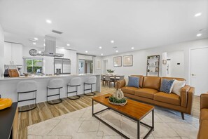 Una cocina moderna con una isla central, taburetes de bar, un comedor con mesa y sillas, un sofá de cuero marrón con cojines estampados y una mesa de centro de madera.