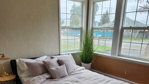 A bedroom with a large bed, a potted plant, and a view of the street through two windows. - Cute Parkside House (Grants Pass)