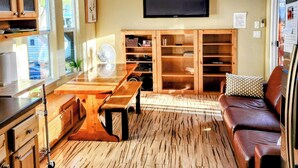 A spacious living area with a wooden floor, a ceiling fan, a flat-screen TV, built-in shelves, a wooden bench, and a leather couch.
