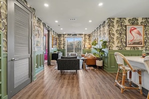 A room with a wooden floor, green walls, a dining area with a table and chairs, and a large window. - Petal & Palm-Rooftop Patio & Gym-Mins to Broadway (Nashville)