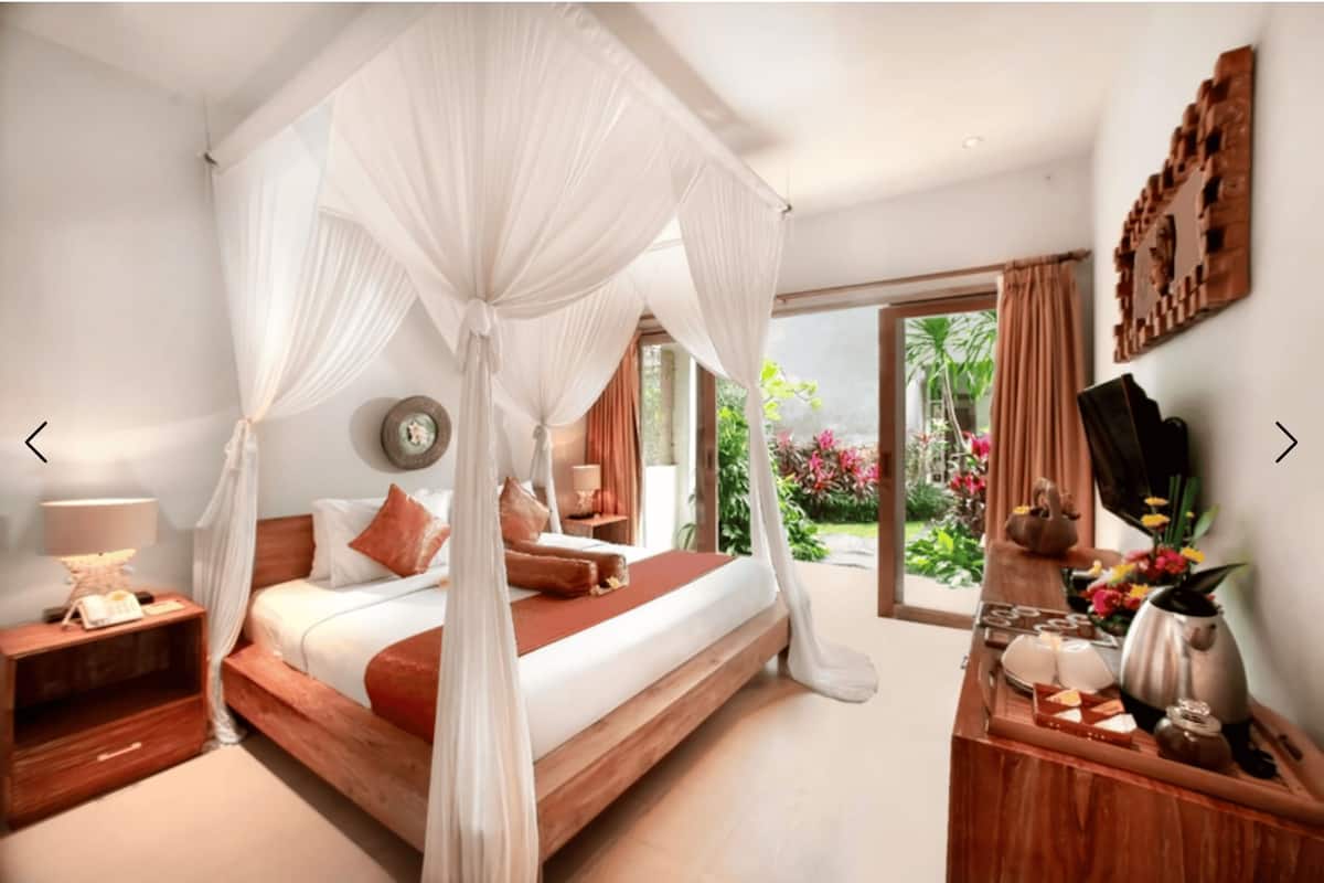A bedroom with a canopy bed, a bedside table with a lamp, a TV, and a coffee table with a teapot and flowers.