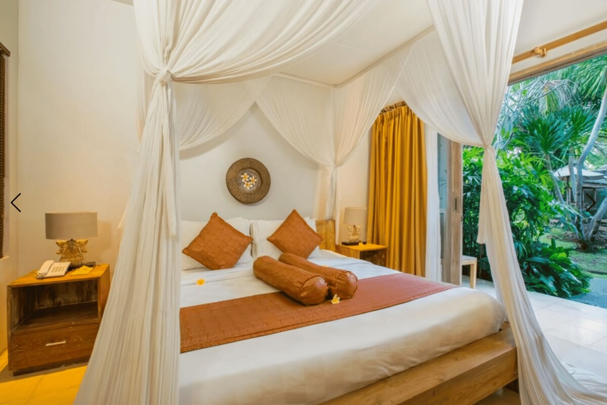 A room with a bed under a canopy, a bedside table with a lamp, and a view of greenery outside.