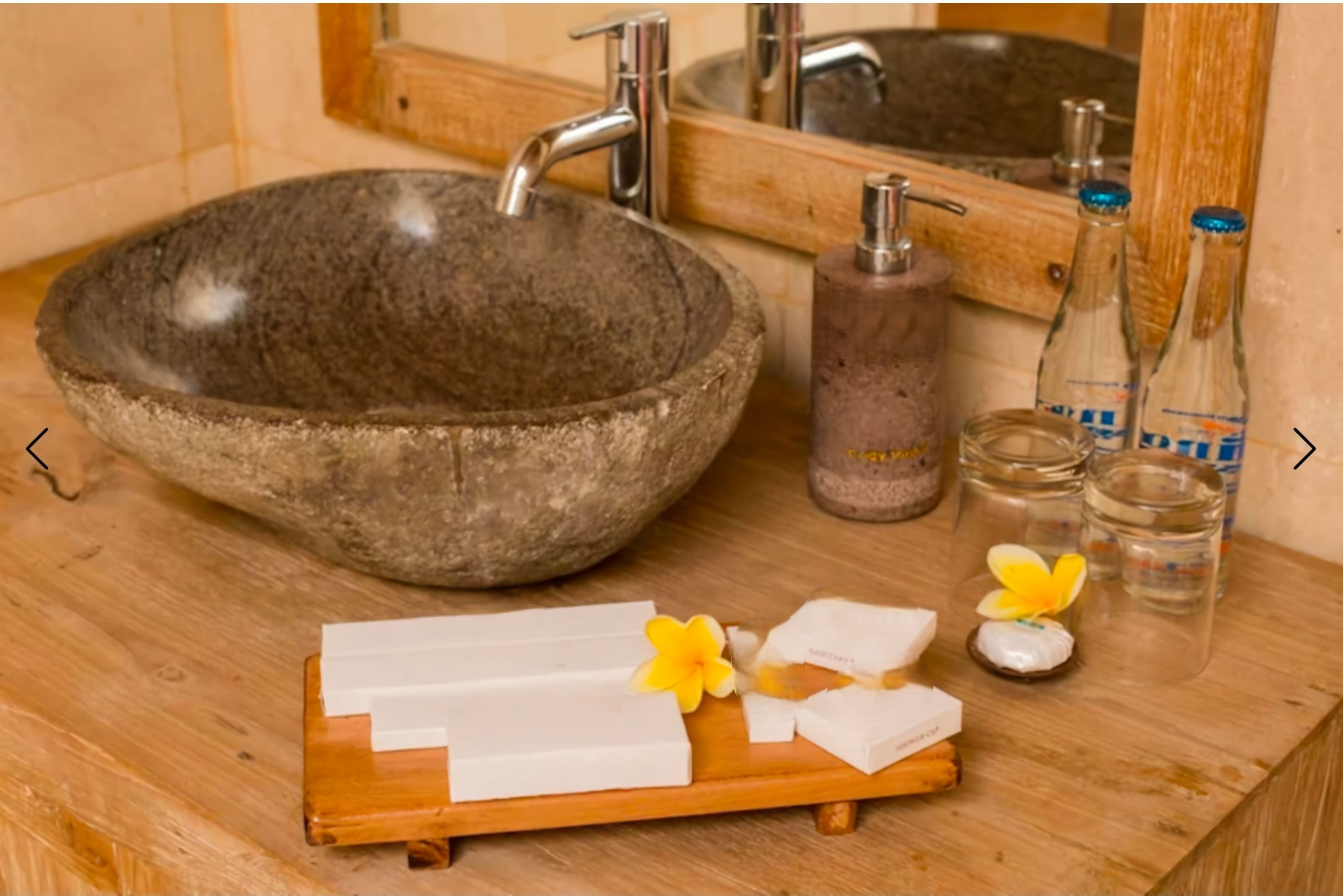 Ein Badezimmer mit einem Steingefäß, einer Holztablett mit Seife und Blumen sowie einem Spiegel mit Flaschen.