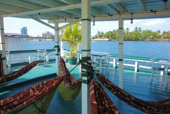 A covered deck with hammocks, a table, chairs, and a view of the water and buildings. - AHHA Floatel Kampot (Santuk)