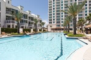 A swimming pool surrounded by lounge chairs and palm trees, with modern buildings in the background. - Sapphire #704 | Beachfront Condo • Gulf & Bay Views • Resort Pool (South Padre Island)