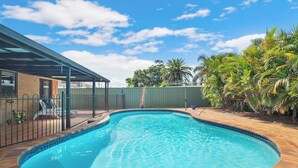 A swimming pool with a wooden deck, surrounded by a fence and palm trees.