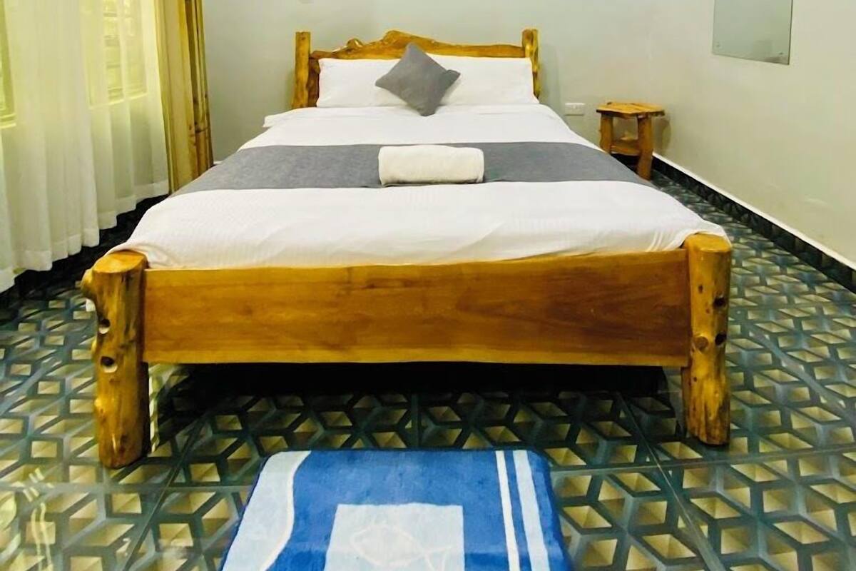 A single bed with a wooden frame, a white mattress, and a gray pillow. There is a small wooden stool beside the bed. The room has a tiled floor with a geometric pattern.