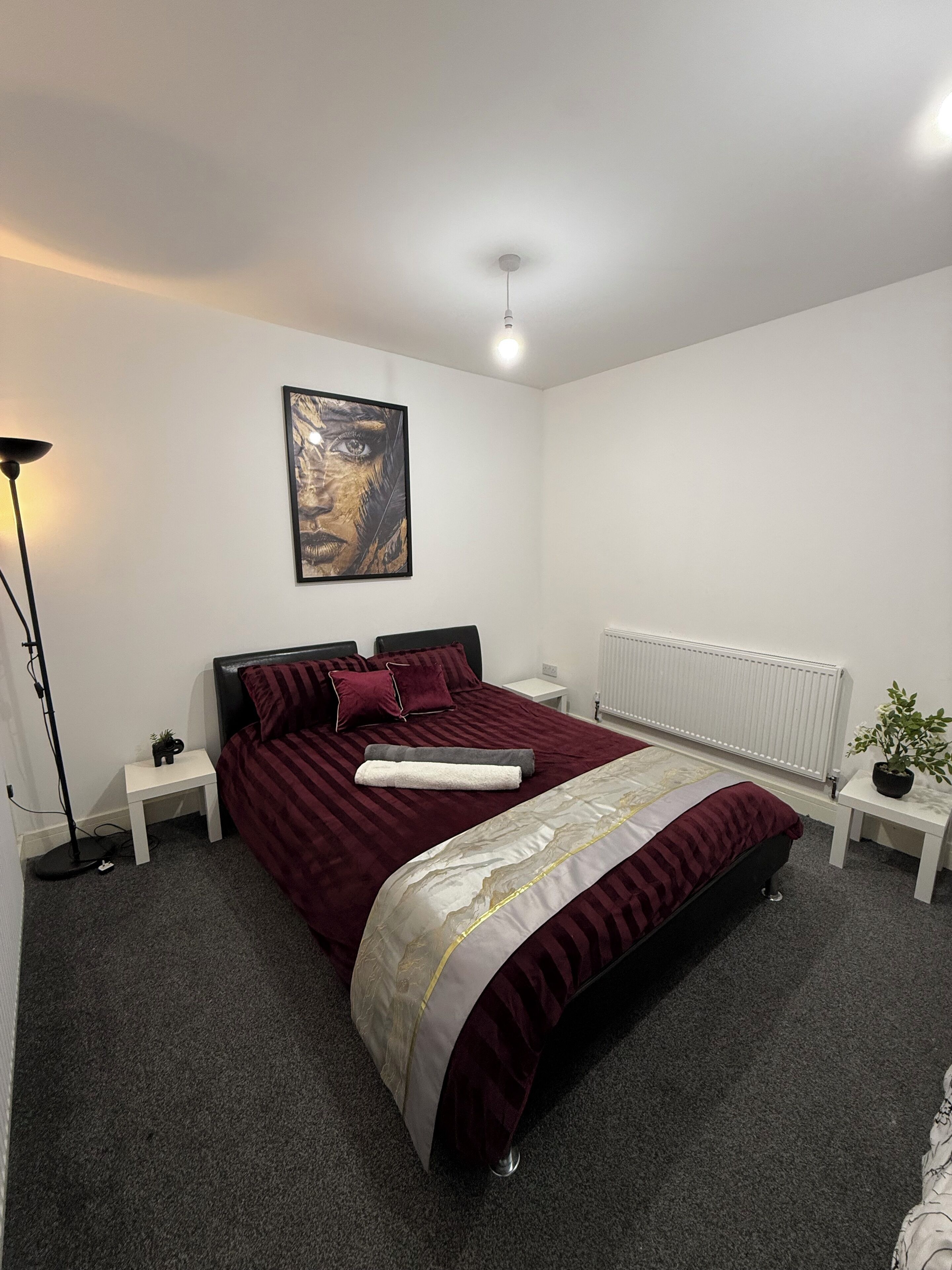 Un lit bien fait, recouvert d’une couvre-lit rouge foncé, agrémenté d’une touche de doré et de blanc. Une petite table de chevet, sur laquelle se trouvent une lampe et une plante en pot, est placée à côté du lit. Un tableau encadré est accroché au mur, au-dessus du lit.