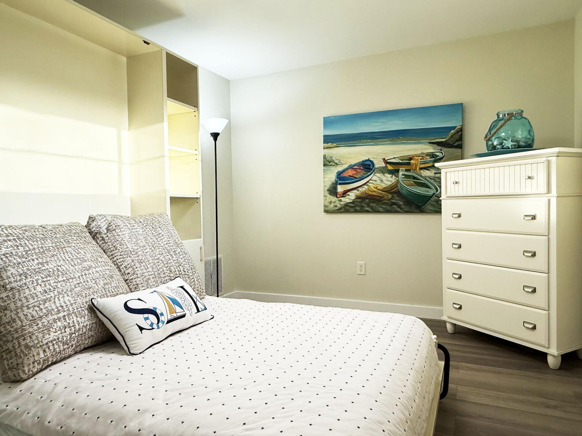 Un dormitorio con cama, cómoda, cuadro de barcos en la playa y una lámpara.