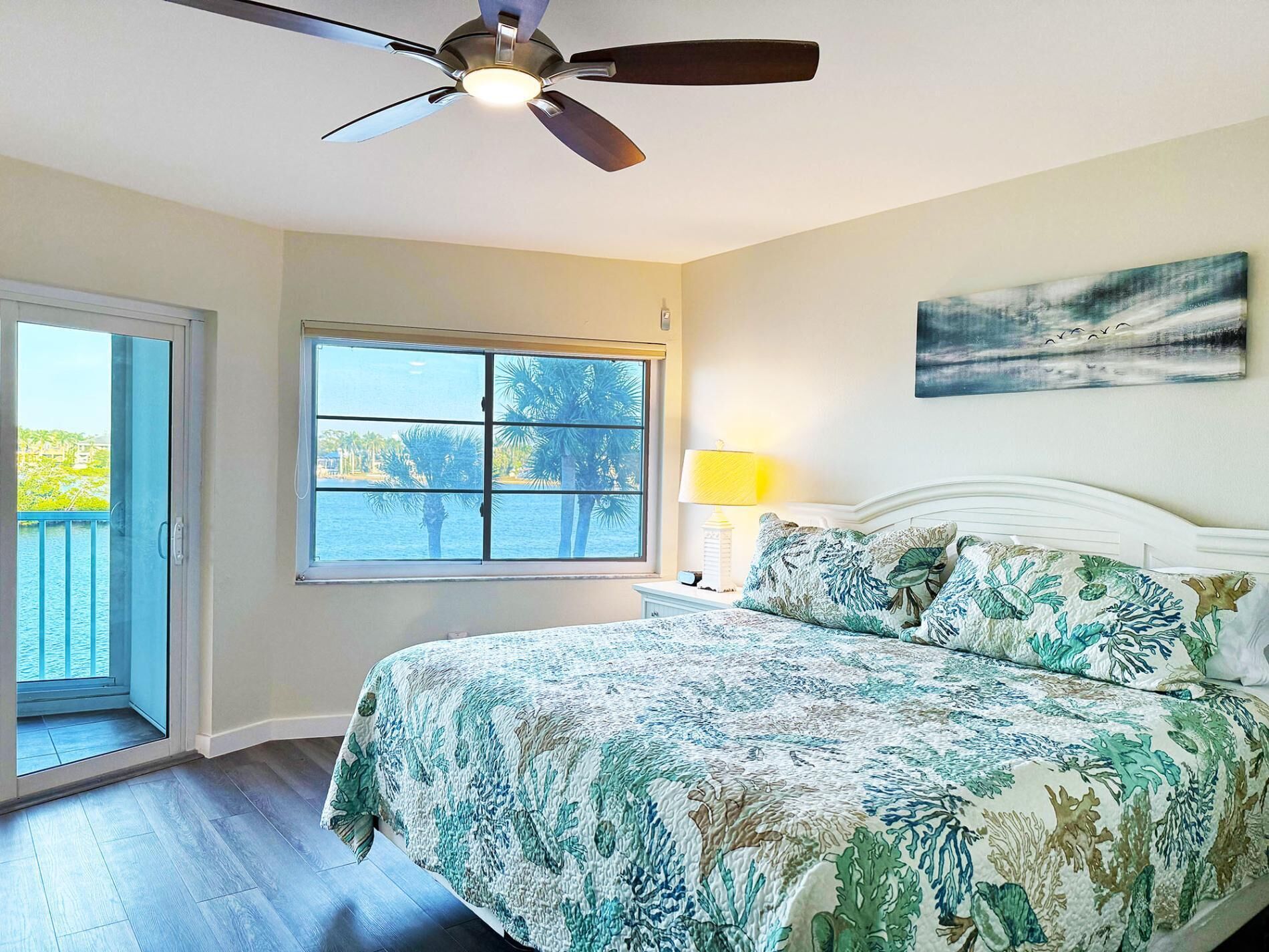 Une chambre avec un lit, un ventilateur de plafond, une fenêtre donnant sur des palmiers et l’eau, et un tableau accroché au mur.