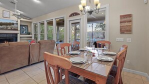 Un coin repas avec une table en bois dressée pour quatre personnes, entourée de chaises en bois. Un lustre est suspendu au-dessus de la table, et on aperçoit une cheminée au fond.