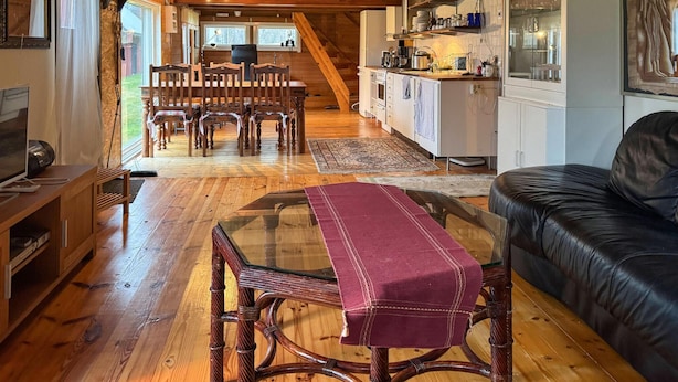 Uma sala de estar acolhedora com piso de madeira, uma mesa de centro de vidro, um sofá de couro e um espaço de jantar com cadeiras e mesa de madeira.