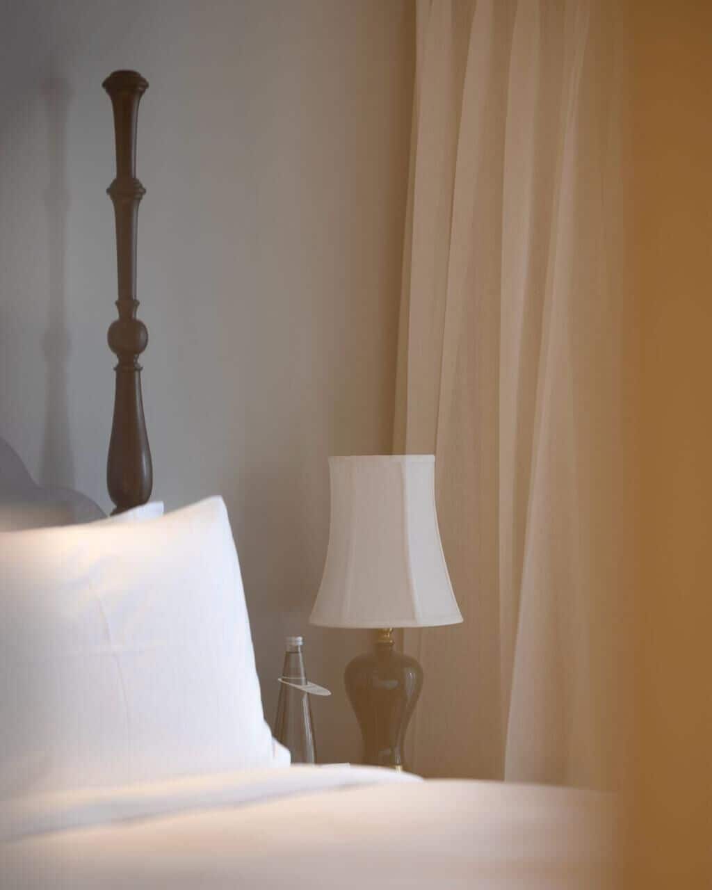 Una cama con un almohadón blanco y un cabecero de madera oscura, una lámpara de noche y una cortina entreabierta.