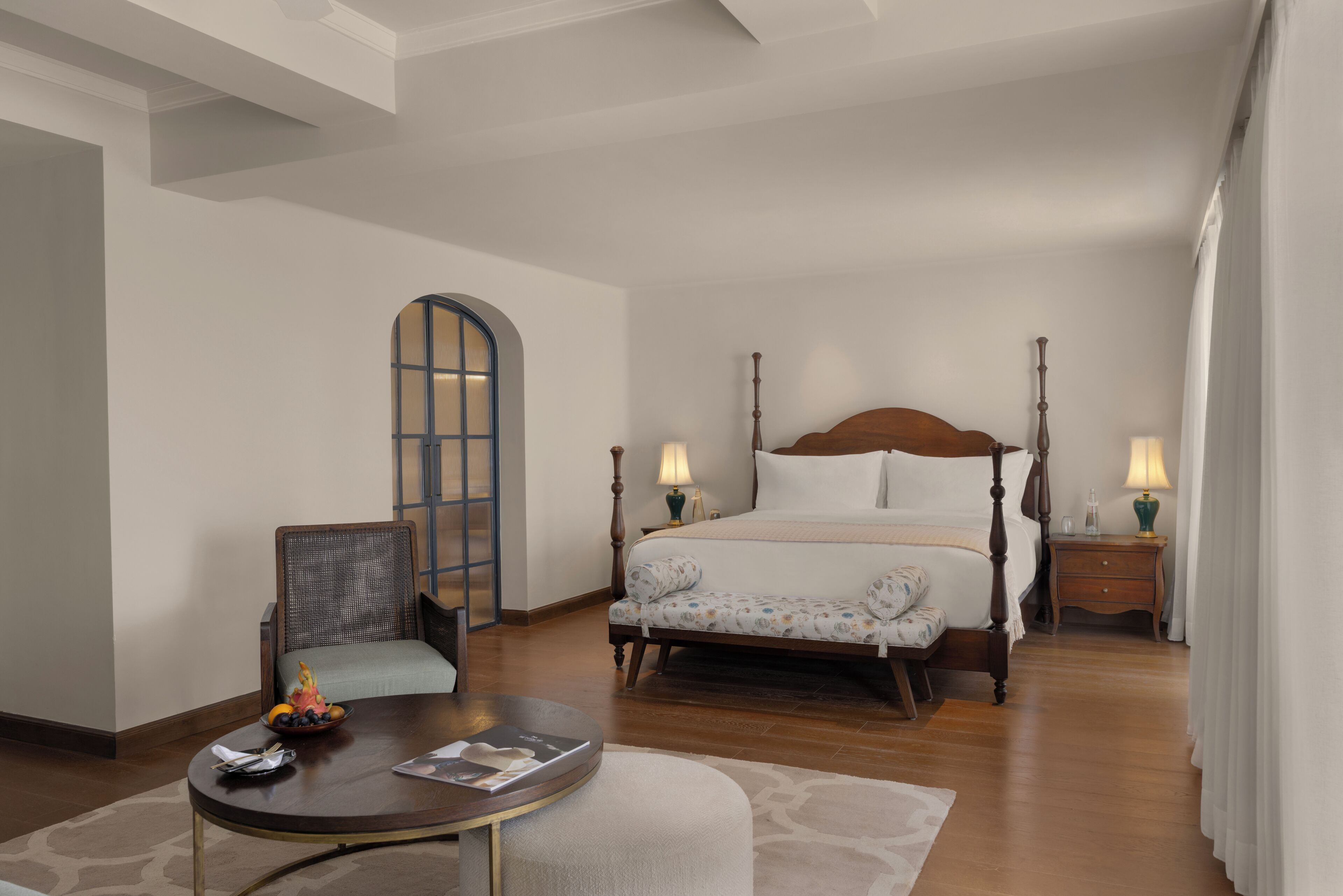 Une chambre à coucher avec un lit à baldaquin, une chaise, une table ronde sur laquelle se trouve un saladier rempli de fruits, et une table de chevet avec une lampe.