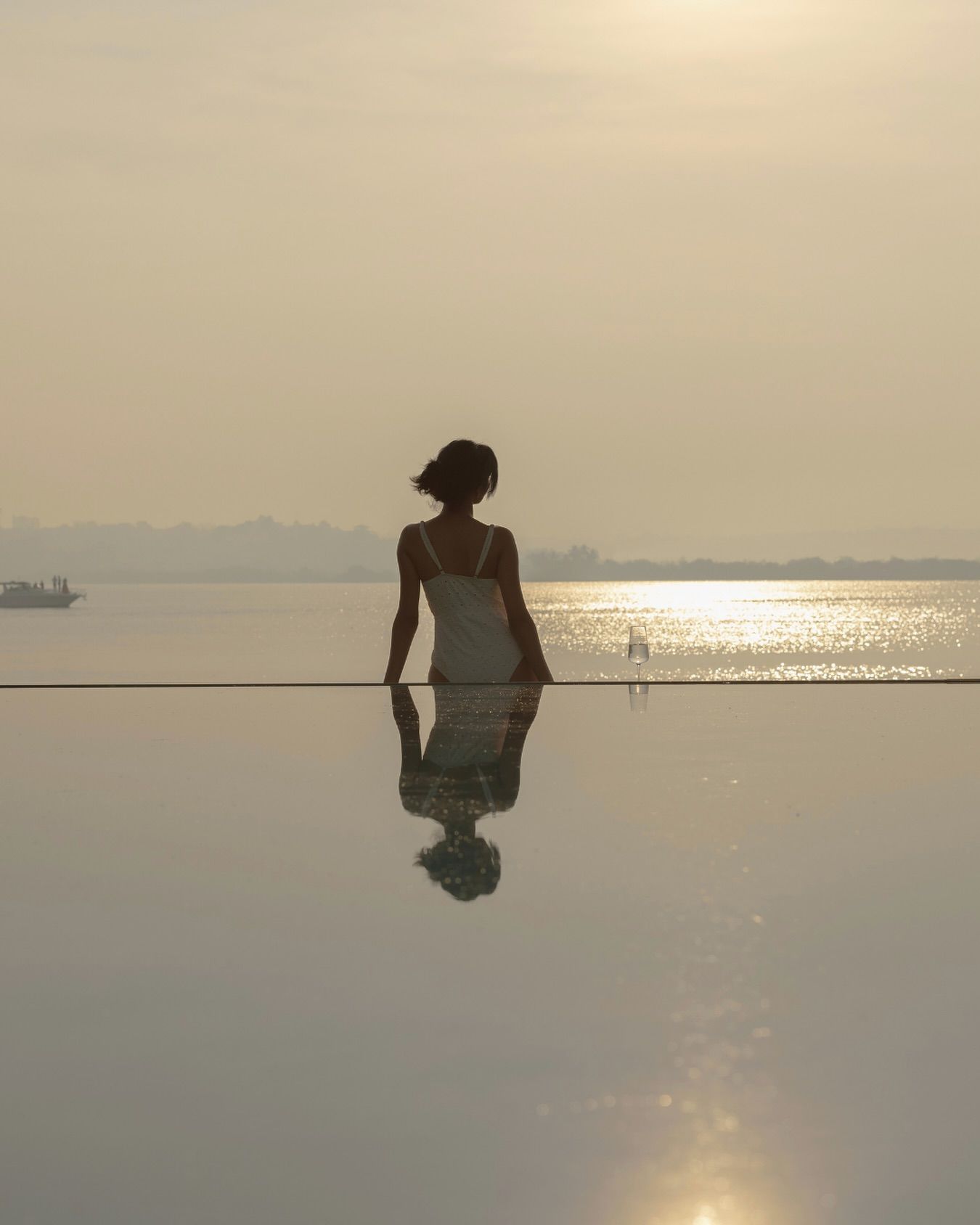 Uma pessoa sentada em frente a uma superfície refletora, com vista para um corpo d'água e uma linha de costa distante, e um barco.