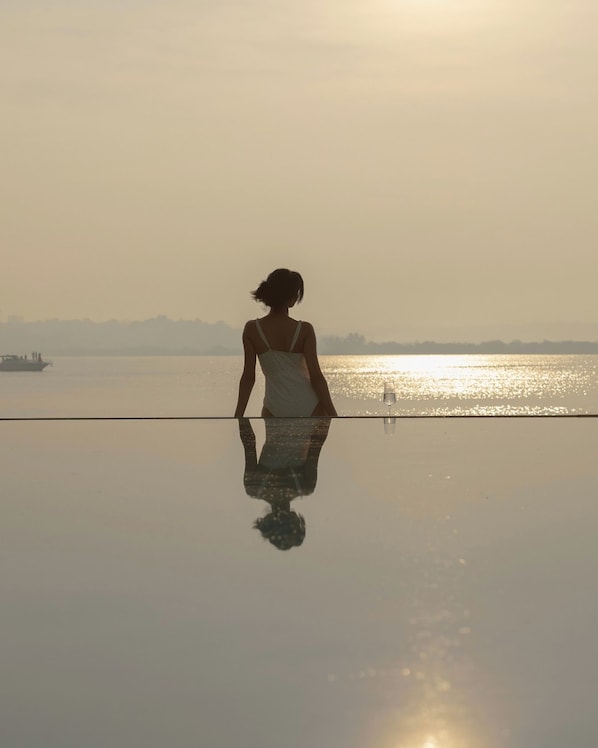 A person seated at a reflective surface overlooking a body of water with a distant shoreline and a boat. - The Postcard on The Mandovi River (Alto de Porvorim)