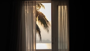 A room with a table and chairs, a view of the sea through a window, and a palm tree outside. - The Postcard on The Mandovi River (Alto de Porvorim)