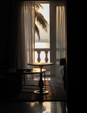 Ein Zimmer mit Tisch und Stühlen, Meerblick durch ein Fenster und eine Palme draußen.